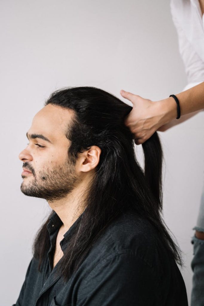 person-parting-the-bearded-man-s-hair-5234227 Profile of a man with long hair being styled into a ponytail.
